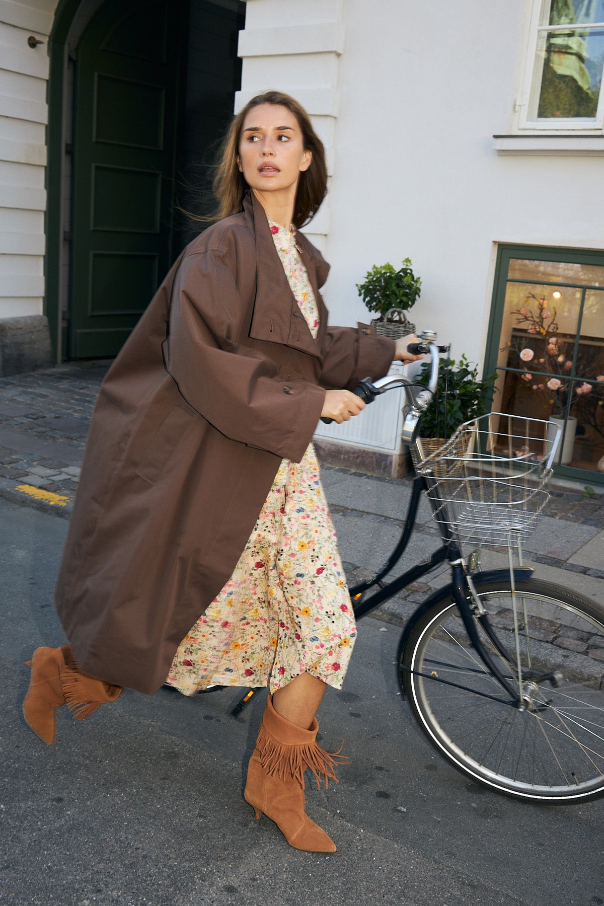 Woman walking her bike on a street in Copenhagen, wearing a long brown coat over a floral dress with knee-high heeled boots.