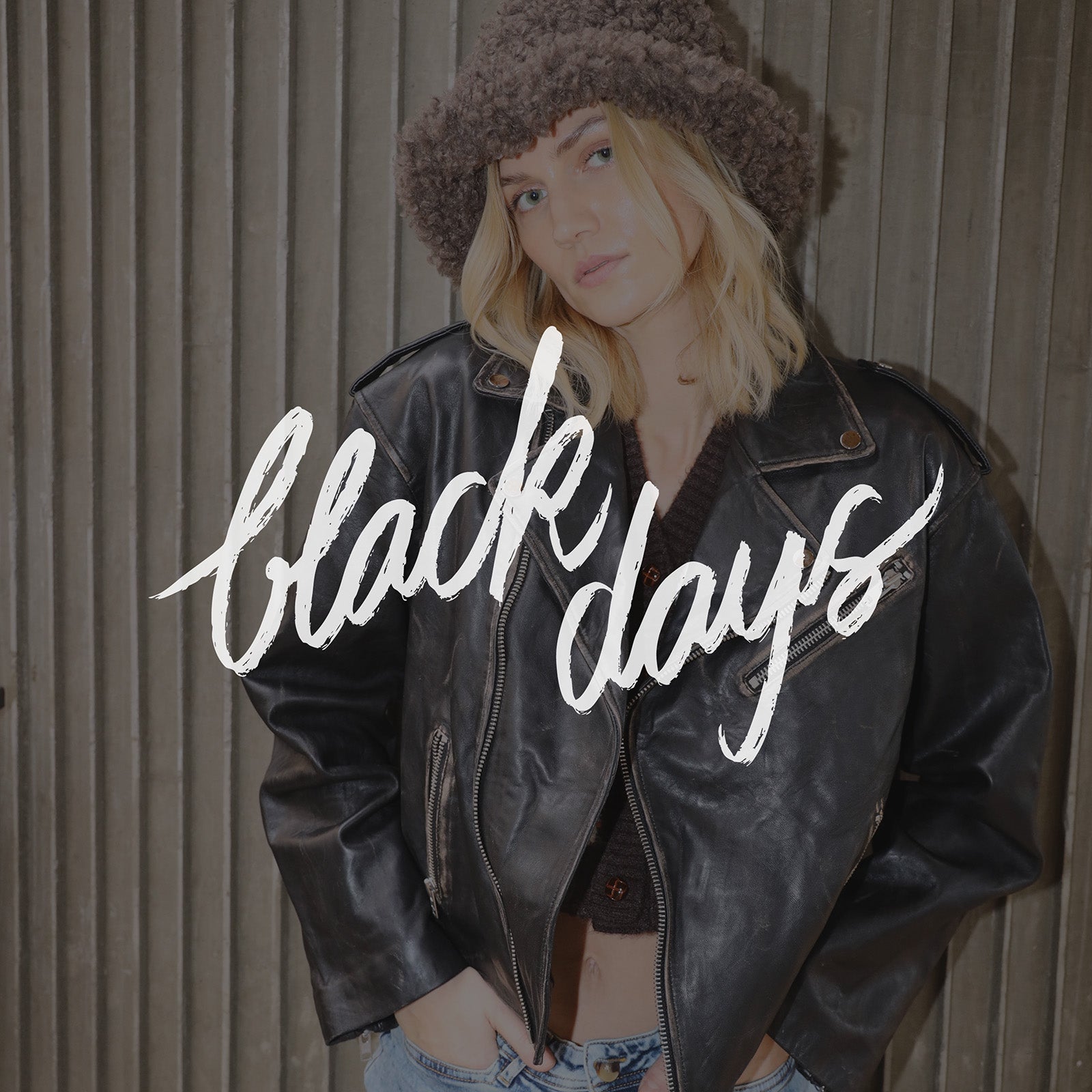 Woman wearing a black leather jacket and a textured bucket hat, standing in front of a ribbed wall. The text black days appears across the image.