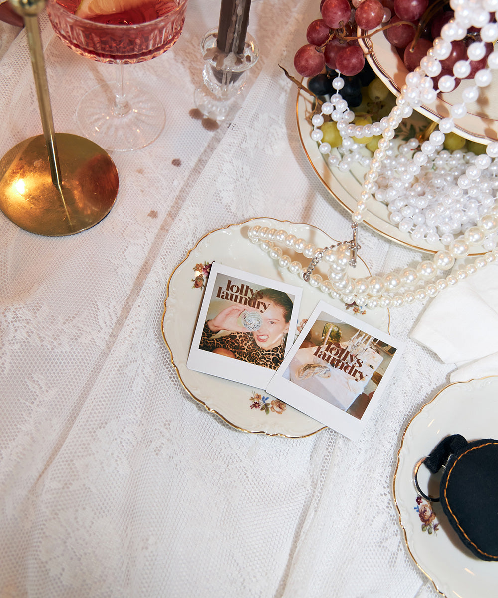 Table setting with a white lace tablecloth, pearl necklaces, grapes, vintage plates, pink glasses and small Lollys Laundry photographs.