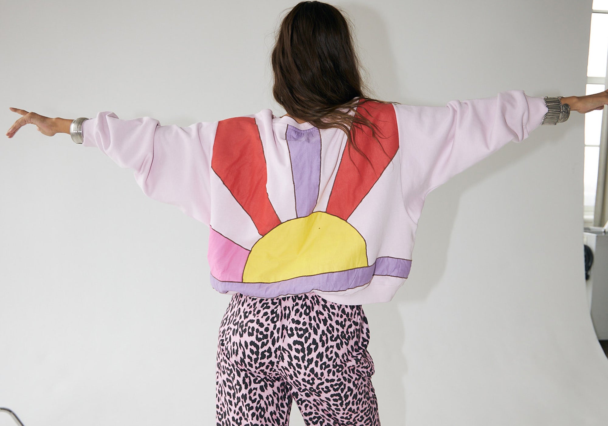 Model wearing a light pink sweatshirt with a sunrise motif in red, purple, and yellow tones on the back, styled with pink leopard-print trousers from Lollys Laundry.