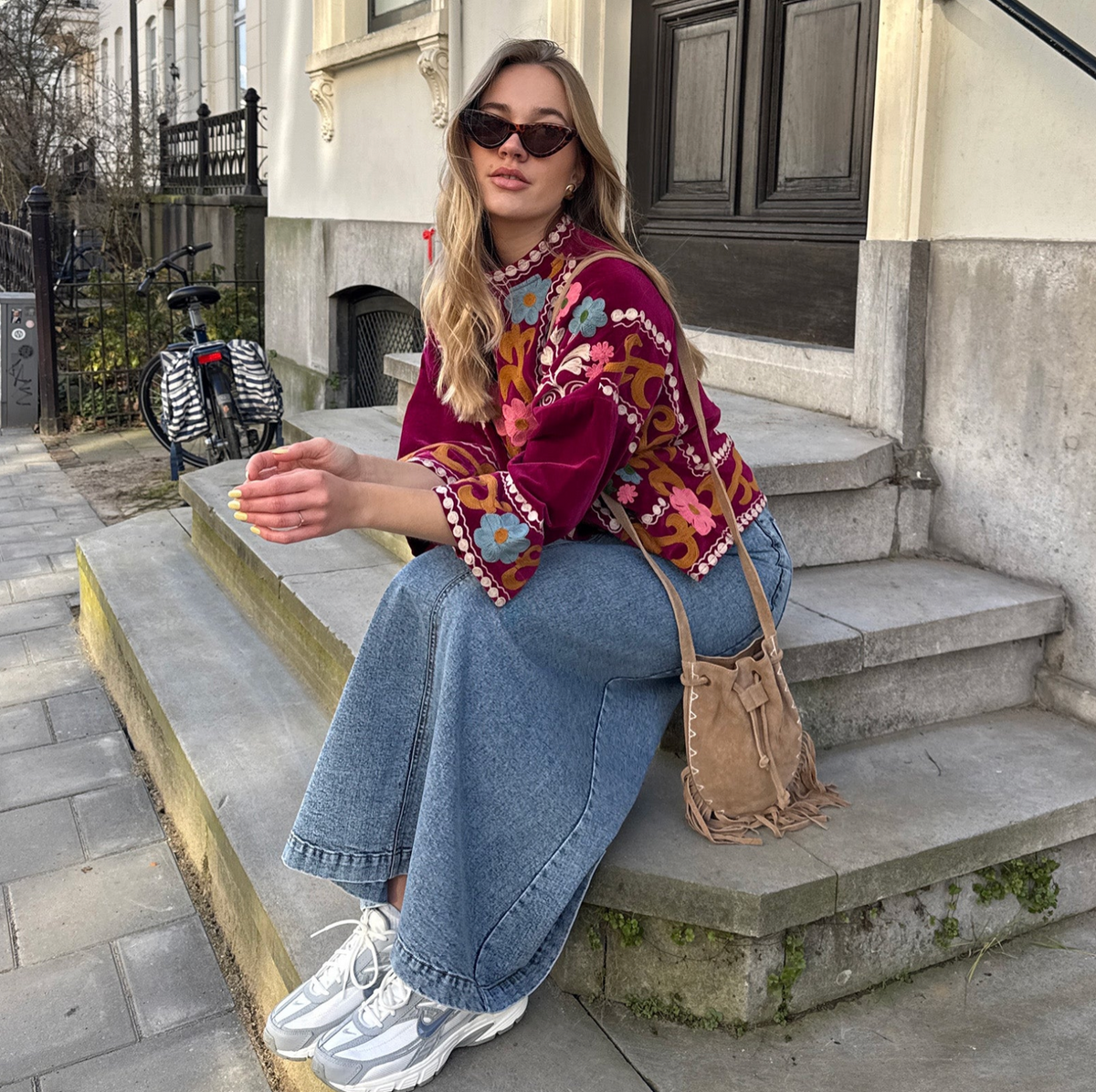 Woman wearing a burgundy jacket from Lollys Laundry with colourful floral embroidery and decorative trims, styled with wide-leg jeans, trainers and a brown shoulder bag.