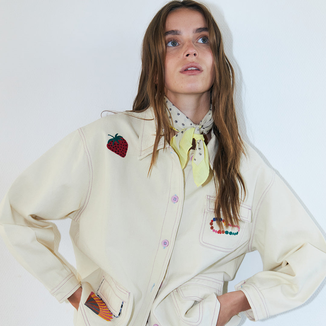 Woman wearing a light cream jacket with playful embroidered details and colorful stitching, styled with a soft scarf tied at the neck, creating a modern and expressive look.