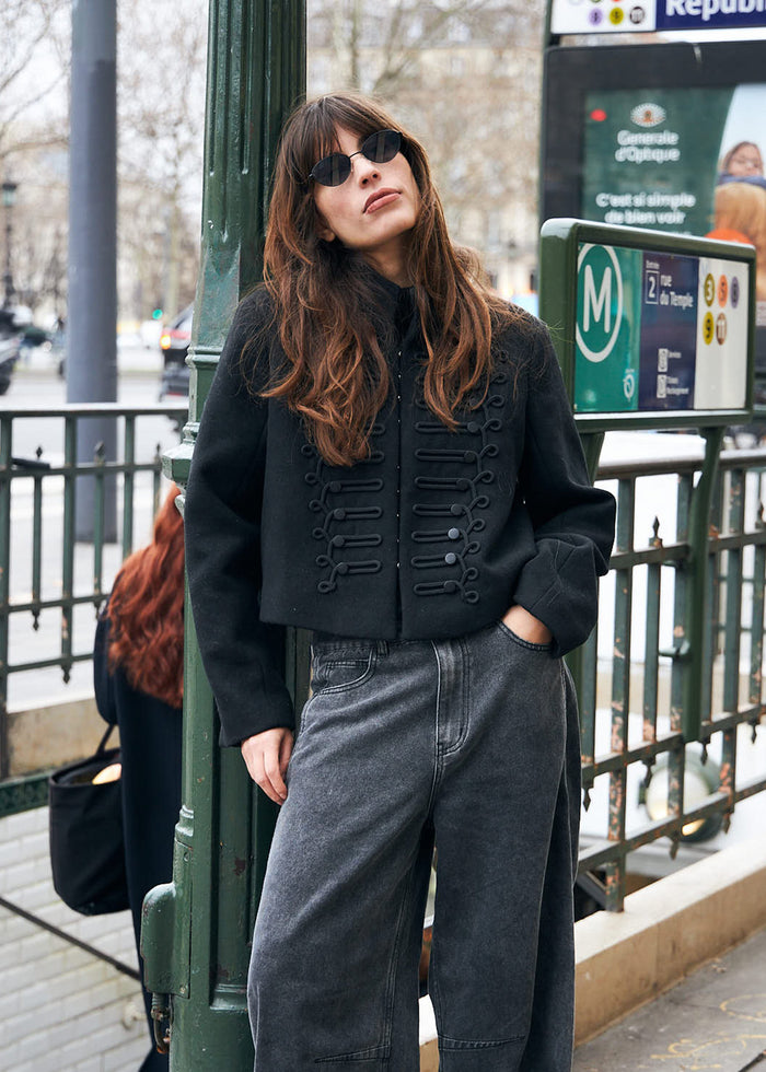 A woman wearing a black croppNapoleon jacket with decorative details, styled with dark grey jeans and black sunglasses, stands on a Parisian Metro platform next to a green pole.
