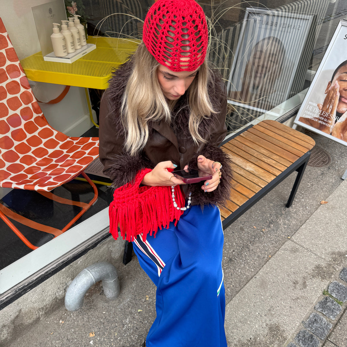 Woman sitting on a bench wearing a brown jacket with fur trims, blue trousers with stripe details and a red crochet hat, with a red fringed bag on her lap.