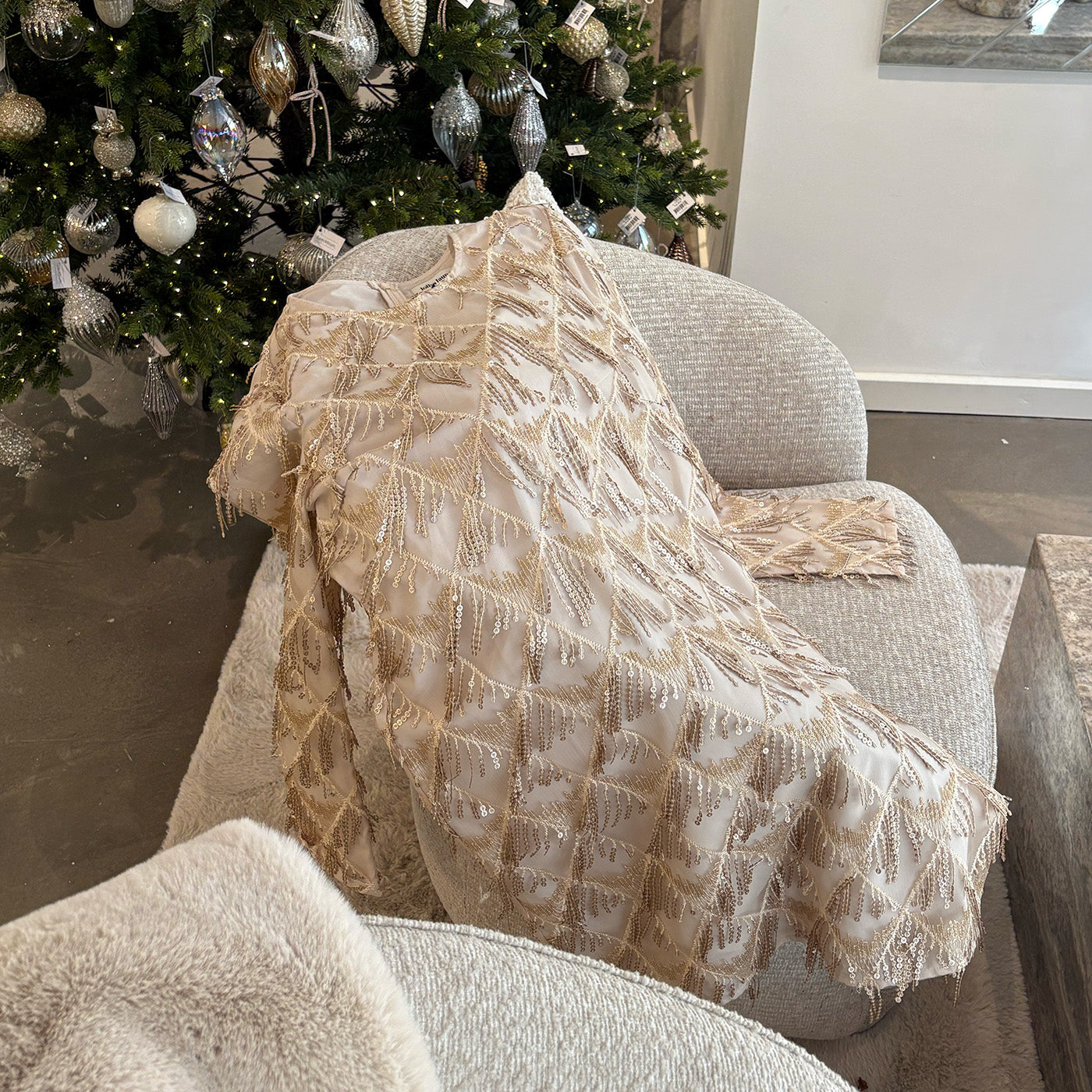 A champagne toned dress with gold beaded details is draped over a light grey lounge chair in front of a decorated Christmas tree adorned with silver and glass ornaments.