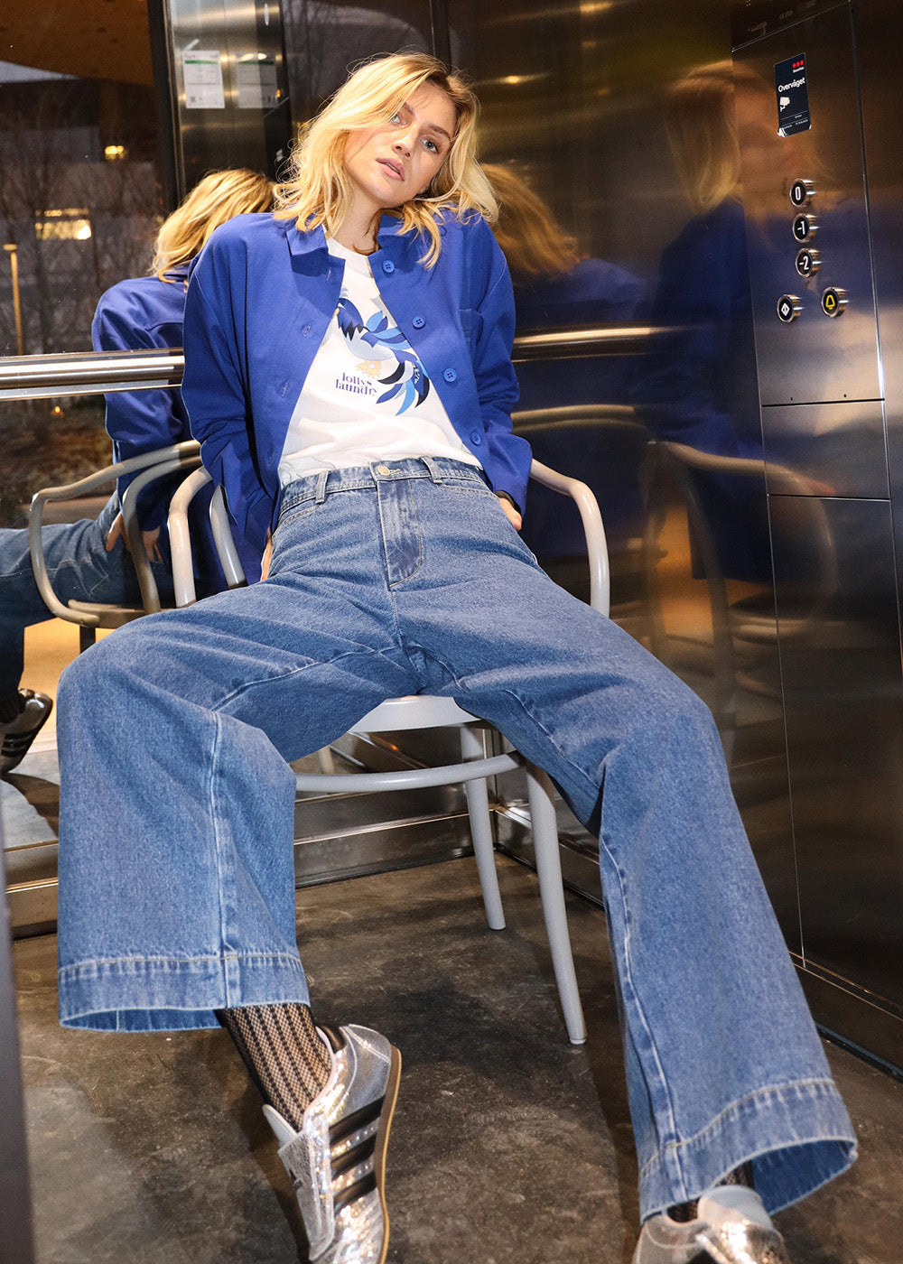 Model sitting on a chair in an elevator with jeans, t-shirt with lollys print and a cool blue jacket