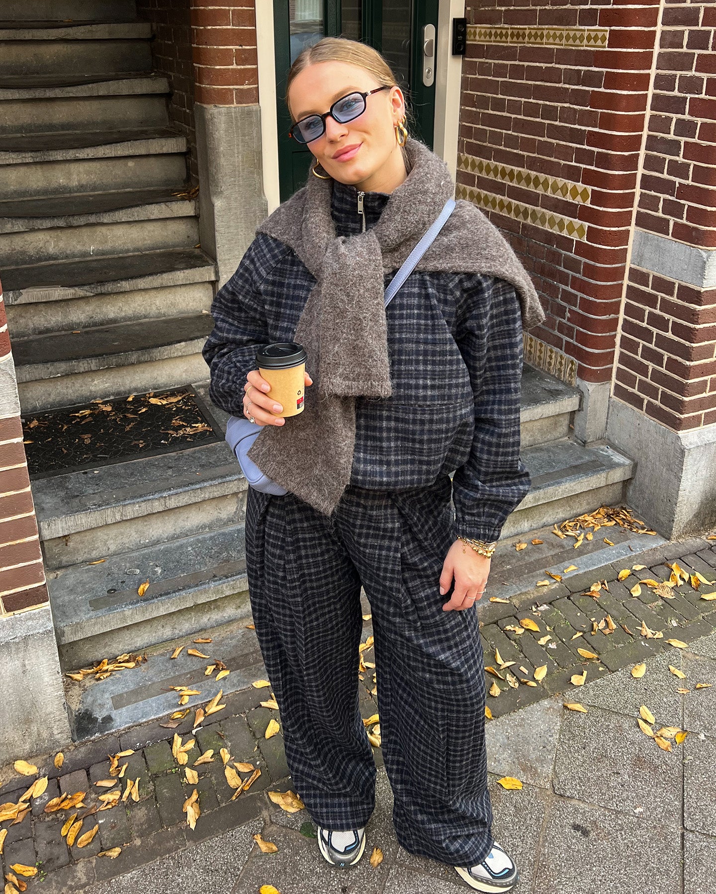 Person in plaid outfit in navy holding a coffee cup on steps with brick building background