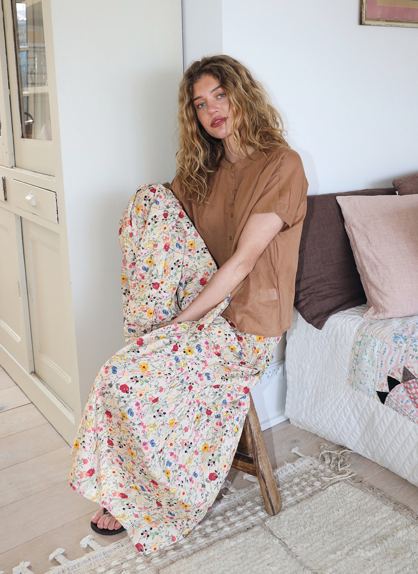 Woman wearing a brown shirt and long skirt with colorful floral print in a light fabric, sitting indoors