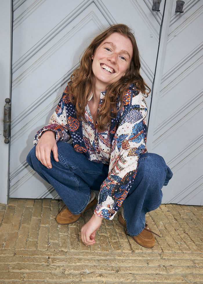 A smiling young woman crouches in front of a gray paneled door wearing a long-sleeve button-up shirt with a colorful abstract pattern in blue, orange, and cream, paired with wide-leg blue jeans and brown suede shoes.