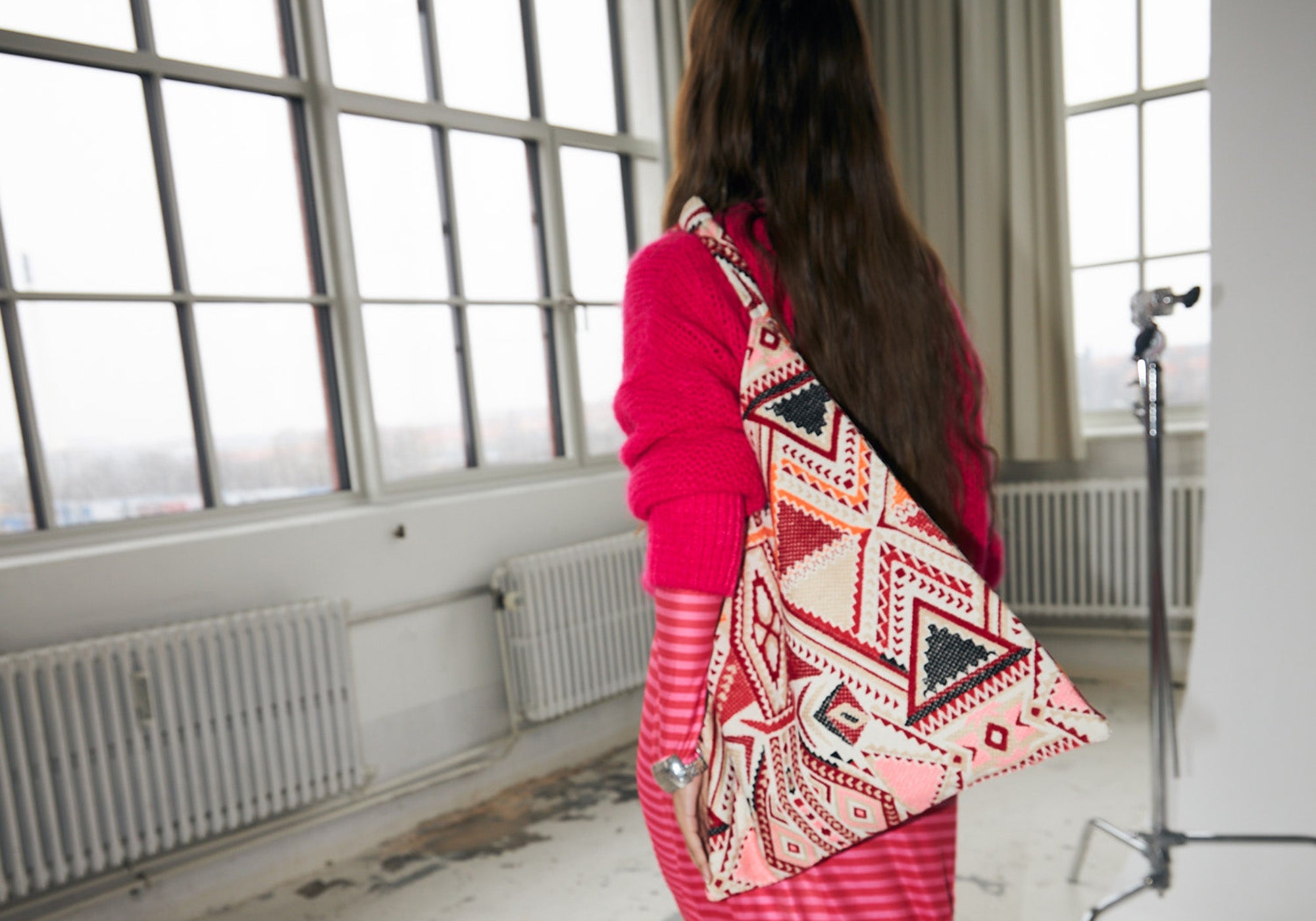 Model wearing a pink knit and striped skirt, carrying a large bag with a colourful geometric pattern in red, orange, and pink tones from Lollys Laundry.