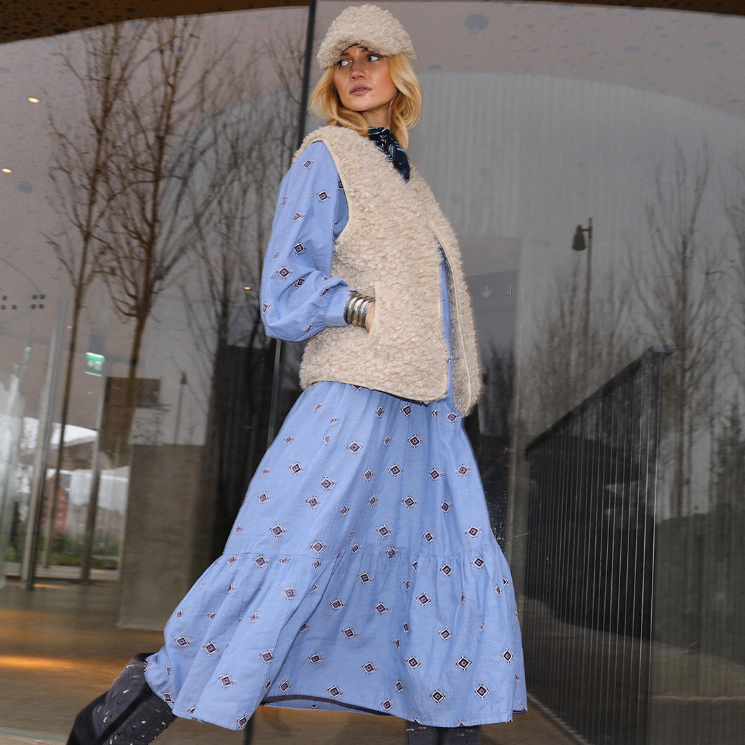 Woman wearing a light blue patterned dress styled with a beige teddy vest and matching hat, photographed outdoors in front of a modern building.