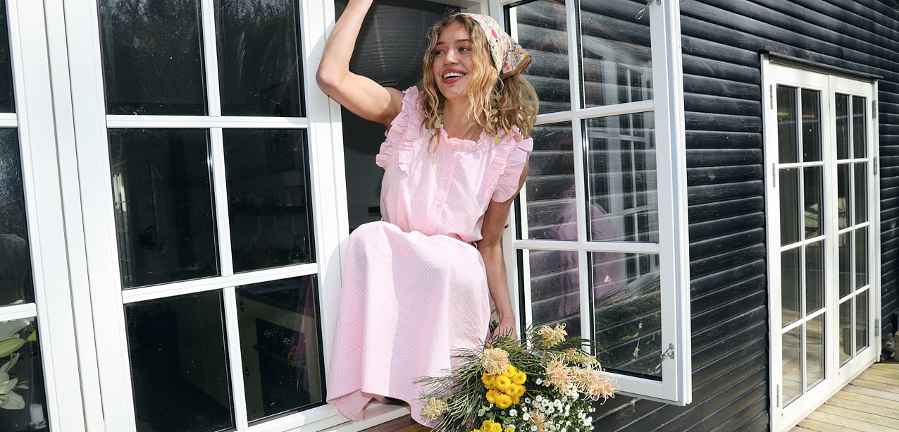 Woman in a light pink dress with ruffle details and a floral scarf sitting in an open window with a bouquet of flowers below.