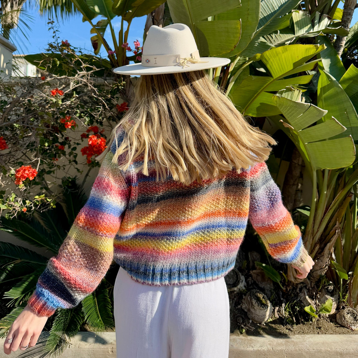 Woman wearing the colourful FairhavenLL knitted jumper from Lollys Laundry with multicoloured stripes in blue, pink, orange and yellow, styled with light trousers and a beige hat.