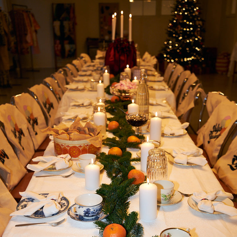 A long dining table set for guests with white tablecloths, blue-and-white porcelain dishes, folded white napkins, and lit white pillar candles. A green pine garland with orange fruits runs down the center, and a decorated Christmas tree glows in the background.