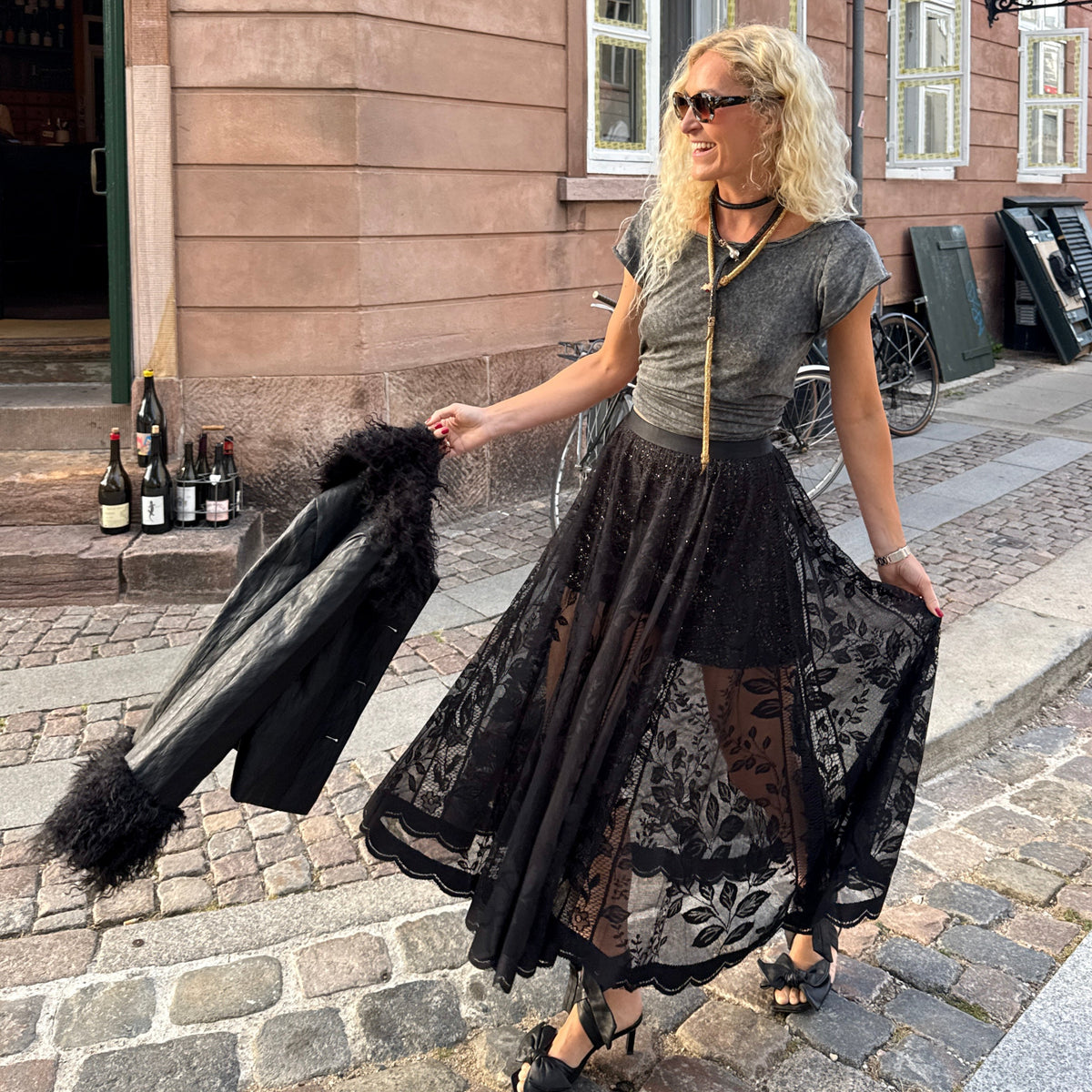 A woman stands on a cobblestone street in front of a brown wooden building, wearing a gray short-sleeve top and a long black lace skirt. She holds a black fur-trimmed jacket in one hand and wears black wedge sandals.