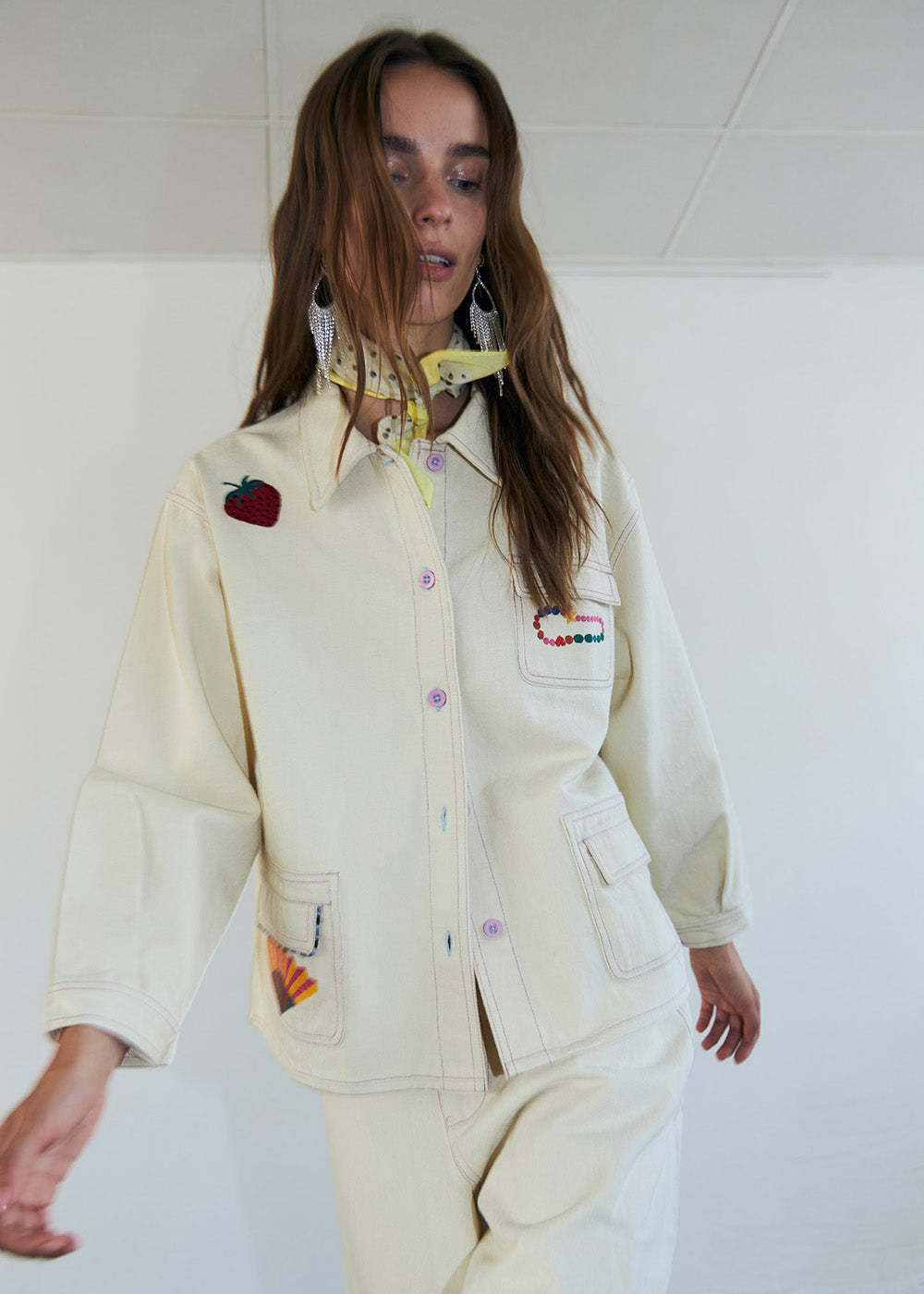 Woman wearing a light jacket with embroidered strawberry and colorful details, styled with matching pants in a minimal studio setting.