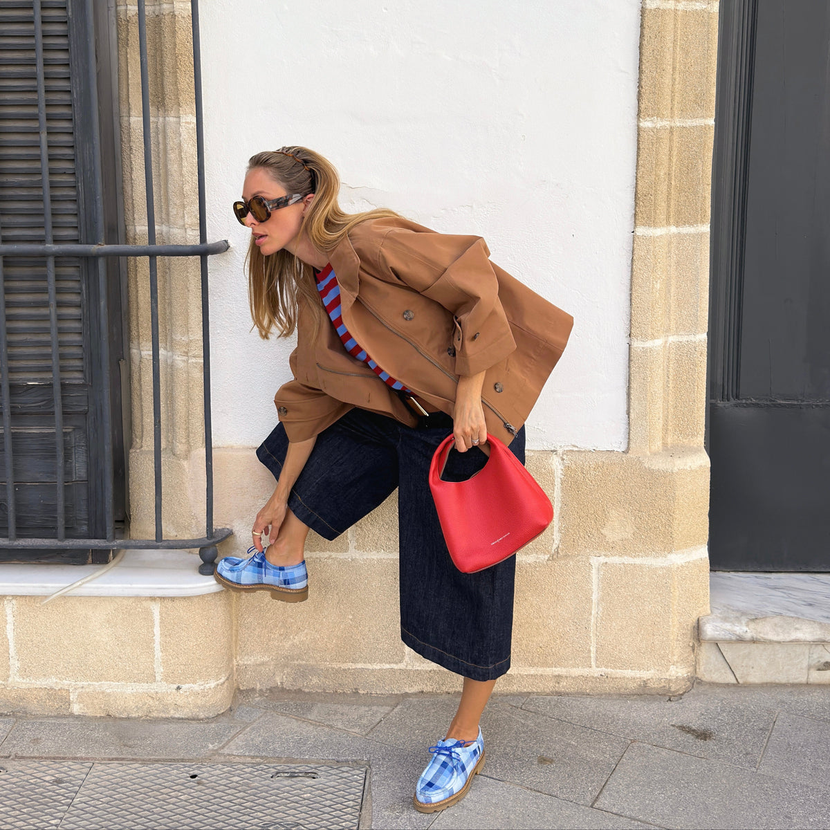 A woman wearing a brown jacket, striped shirt, dark denim shorts, and blue-and-white checkered sneakers poses on a stone ledge next to a white wall, holding a bright red handbag.