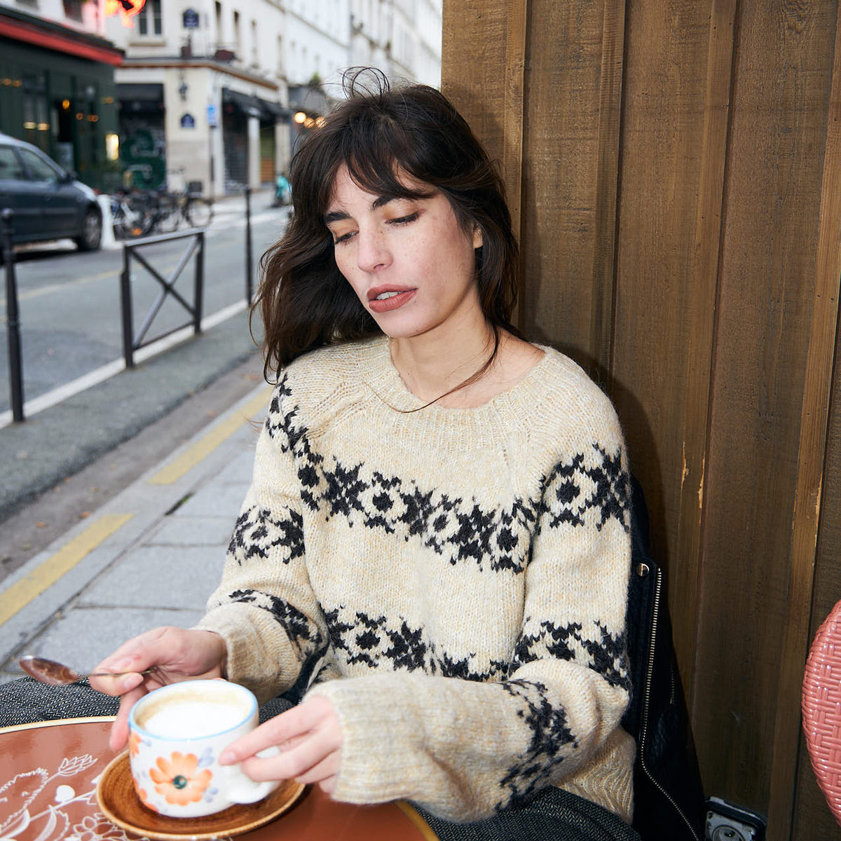 Model wearing a cream Lollys Laundry knit sweater with a dark Nordic pattern, sitting outdoors at a café with a cup of coffee.