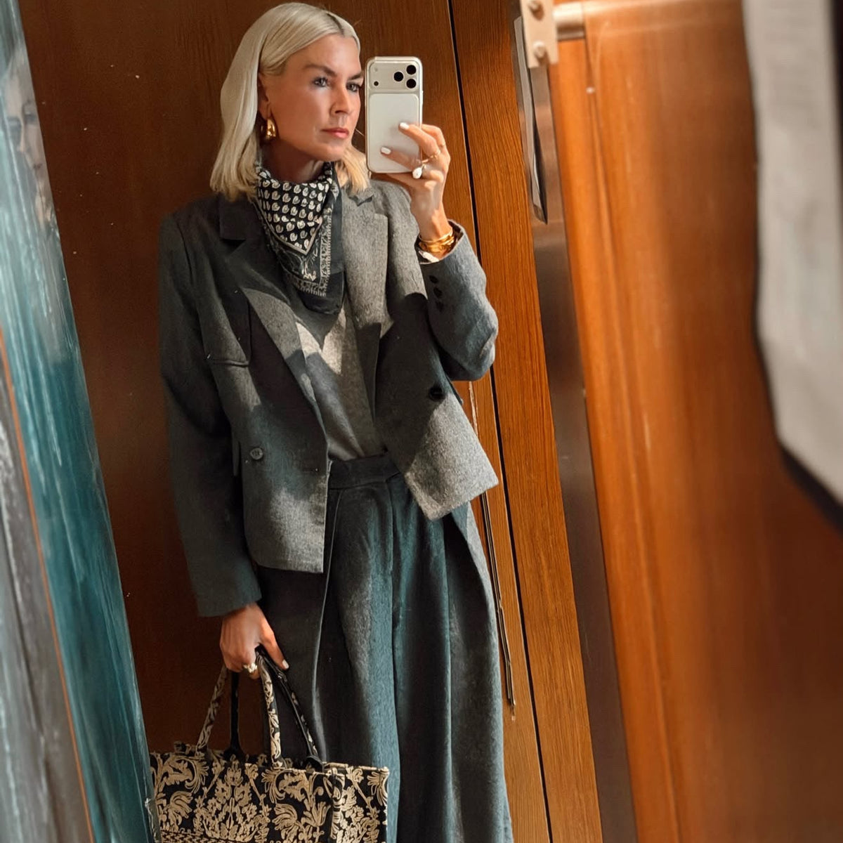 A woman takes a mirror selfie in an indoor setting with wood-paneled walls, wearing a gray blazer, patterned scarf, wide-leg gray pants, and holding a black and beige patterned tote bag.