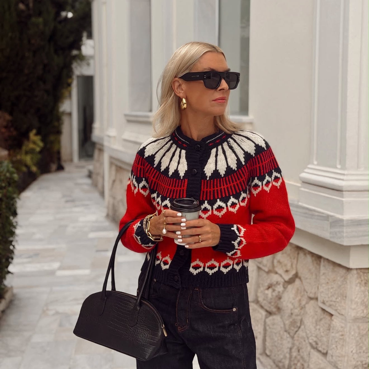 Frau in der ClareLL Strickjacke von Lollys Laundry mit rot-schwarzem und cremefarbenem Muster, kombiniert mit dunkler Jeans, Sonnenbrille und schwarzer Handtasche.