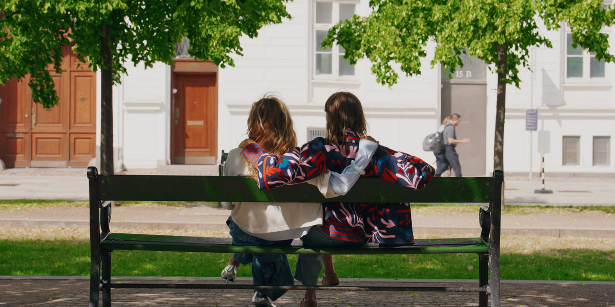 Two women sit with their arms around each other on a bench in Copenhagen, wearing colourful styles from Lollys Laundry.