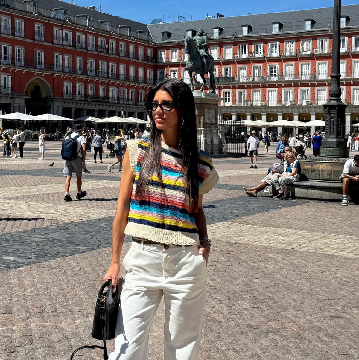 Woman wearing a colourful knitted top with horizontal stripes, styled with white trousers, sunglasses and a black bag in a sunny city square.