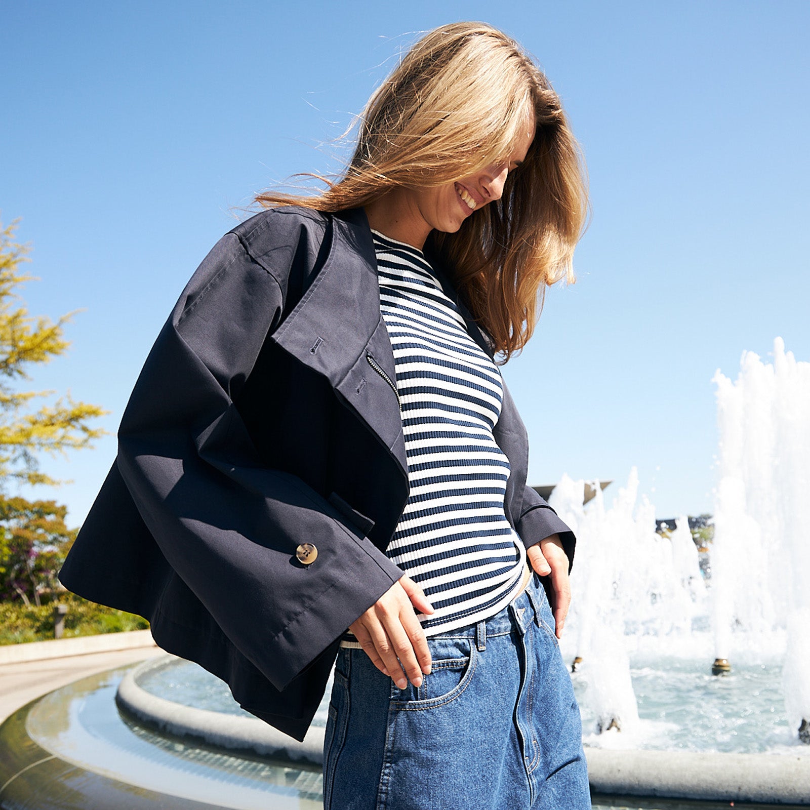 Woman wearing a navy jacket, a striped T-shirt and blue jeans, standing by a fountain on a sunny day.