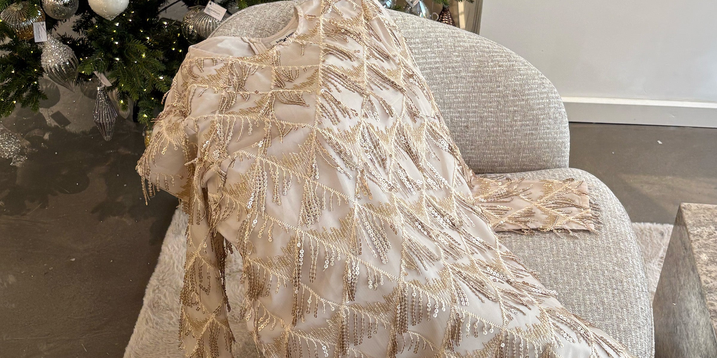 A champagne toned dress with gold beaded details is draped over a light grey lounge chair in front of a decorated Christmas tree adorned with silver and glass ornaments.