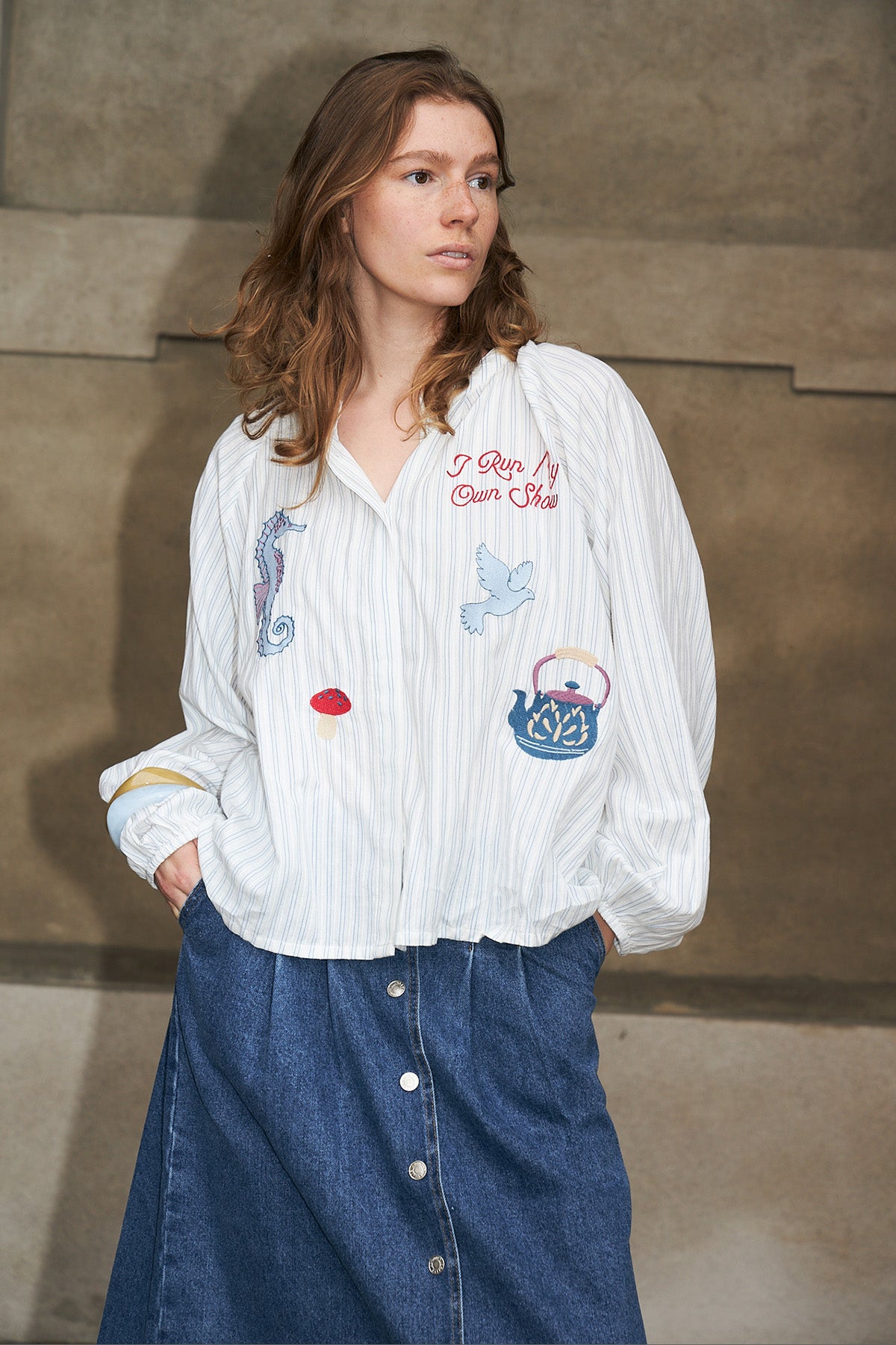 Woman wearing a striped embroidered shirt and a denim skirt, leaning against a wall in Copenhagen.