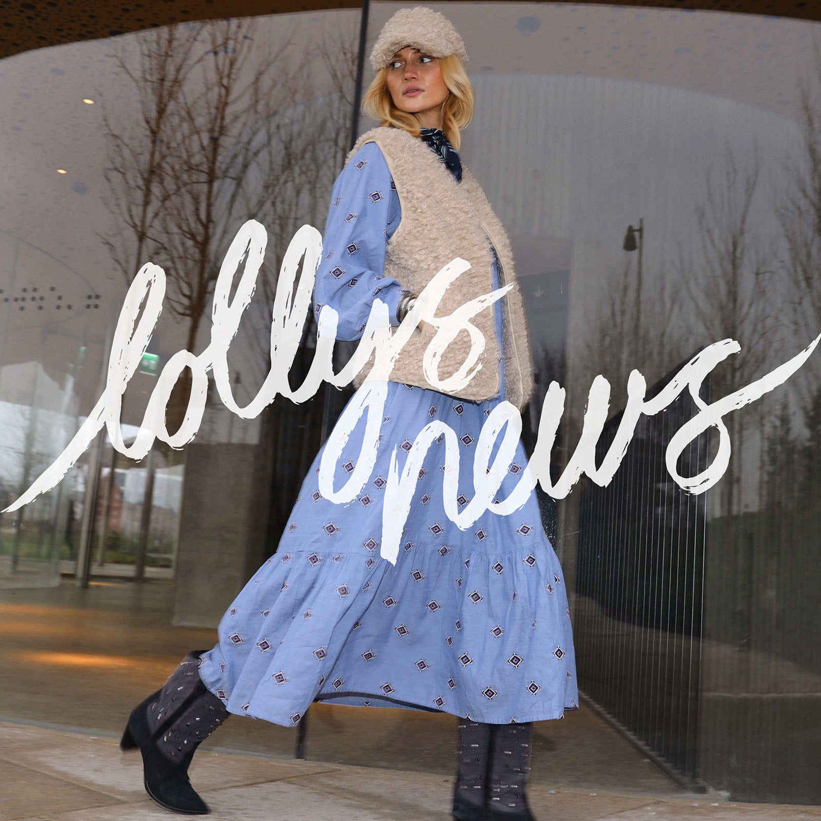 Woman in a blue dress and beige cardigan standing in front of a glass door with 'lollys news' branding.