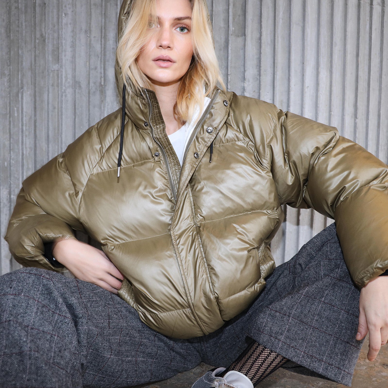 Person wearing a olive green puffer jacket and checked pants sitting on the floor against a neutral background
