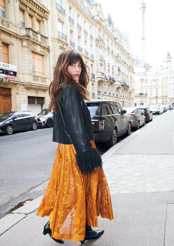 A woman walking on a city street wearing a black faux leather jacket with black fur trim and a long, wide orange patterned skirt.