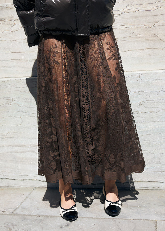A person wearing a long brown sheer lace midi skirt with a floral leaf pattern, styled with a Brown puffer jacket and black-and-white kitten ballerinas standing against a light gray stone wall.