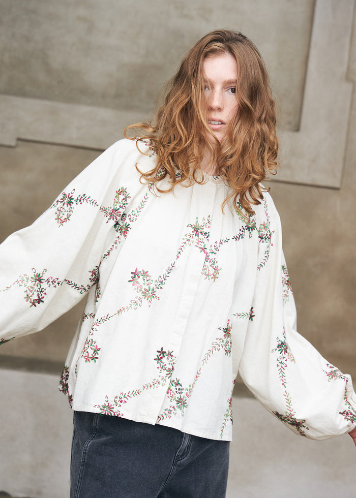 Woman wearing a white blouse with colourful floral embroidery and voluminous sleeves, styled with dark denim jeans. A feminine everyday look with decorative details.