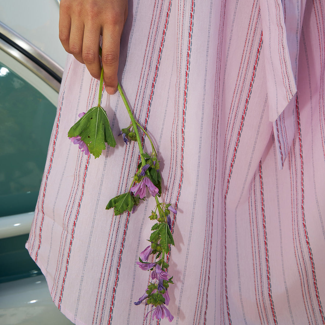 Close up of a striped pink dress with red and blue lines, styled with a hand holding purple flowers.