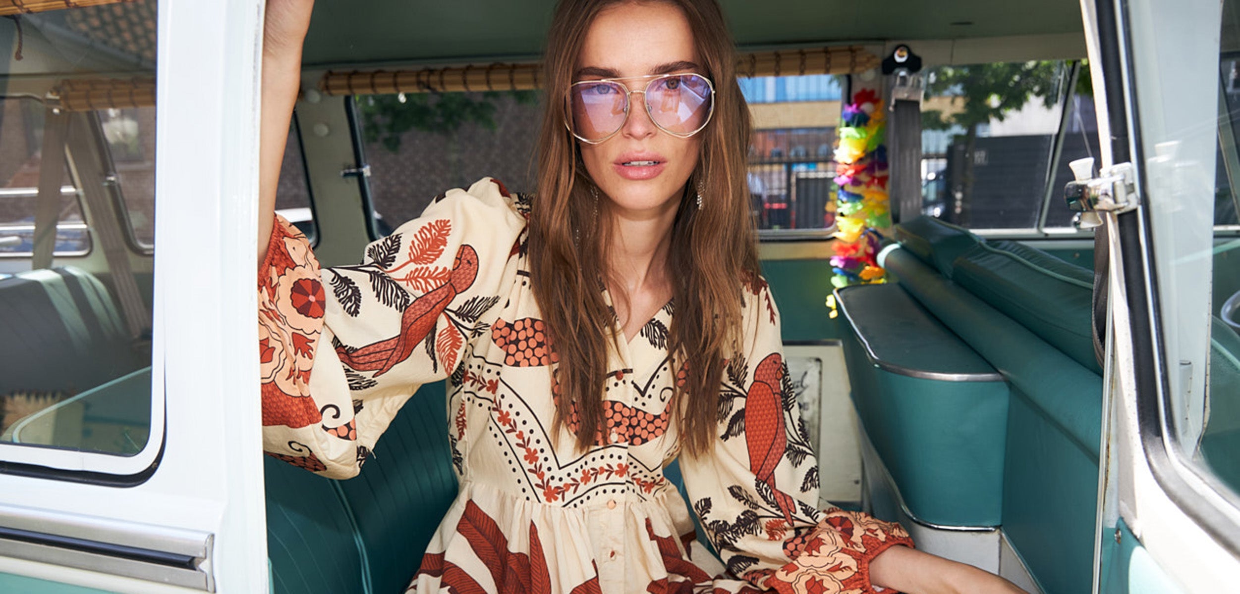 Woman in a long cream dress with a brown botanical print and sunglasses sitting inside a vintage vehicle with colorful interior.