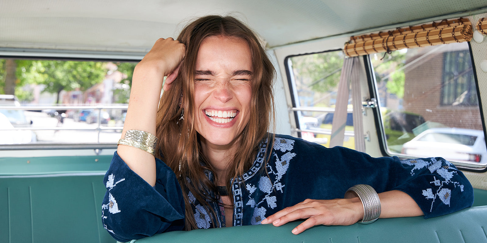Smiling woman sitting inside a vintage camper van wearing a dark blue jacket with light embroidered details. The image captures a relaxed summer mood, effortless elegance, and a bohemian inspired style.