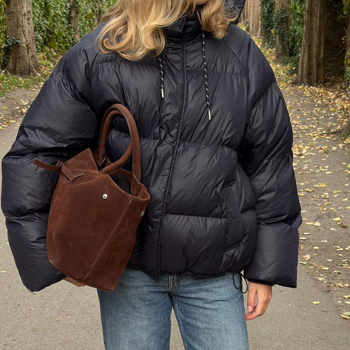 A woman standing outdoors on a paved path wearing a black puffer jacket, blue jeans, and brown shoes, while carrying a brown suede tote bag.