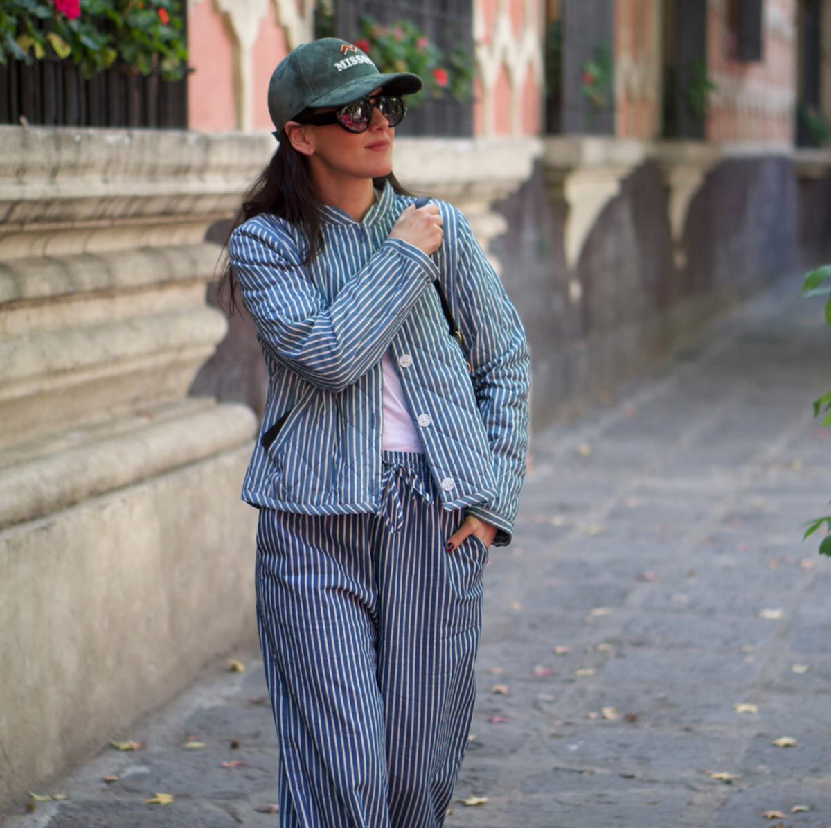 A woman wearing a matching blue and white striped set with a drawstring waist, walking on a stone pathway outdoors. She is also wearing a green cap, sunglasses, and a light pink top underneath.