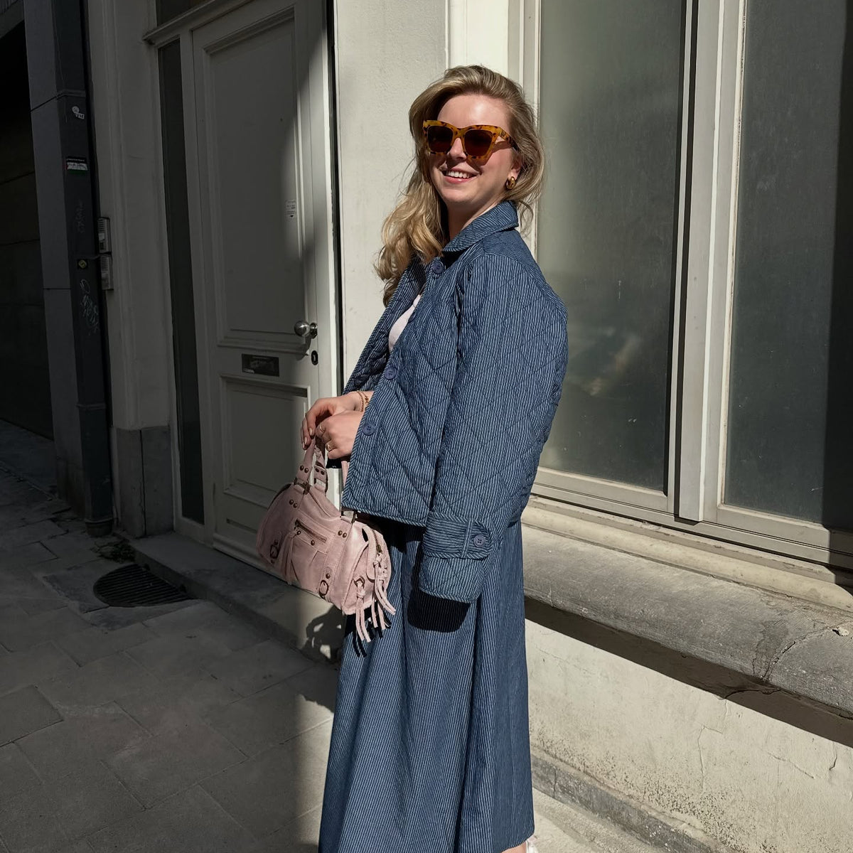 Woman wearing a blue patterned jacket with black contrast details, styled with matching trousers, sunglasses and a pink handbag.