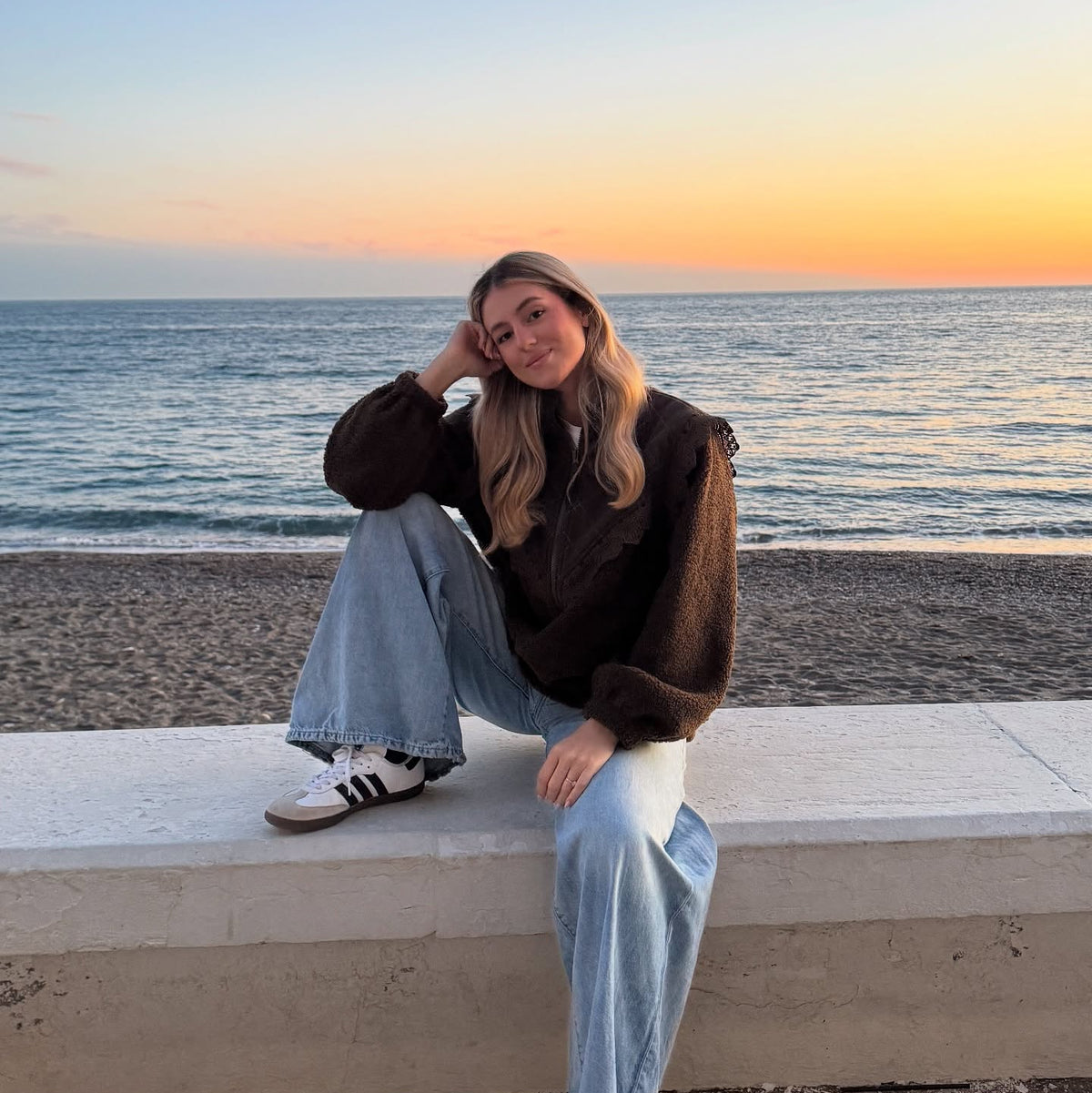 Woman wearing a dark brown textured teddy jacket, light blue jeans and trainers, sitting by the beach at sunset.