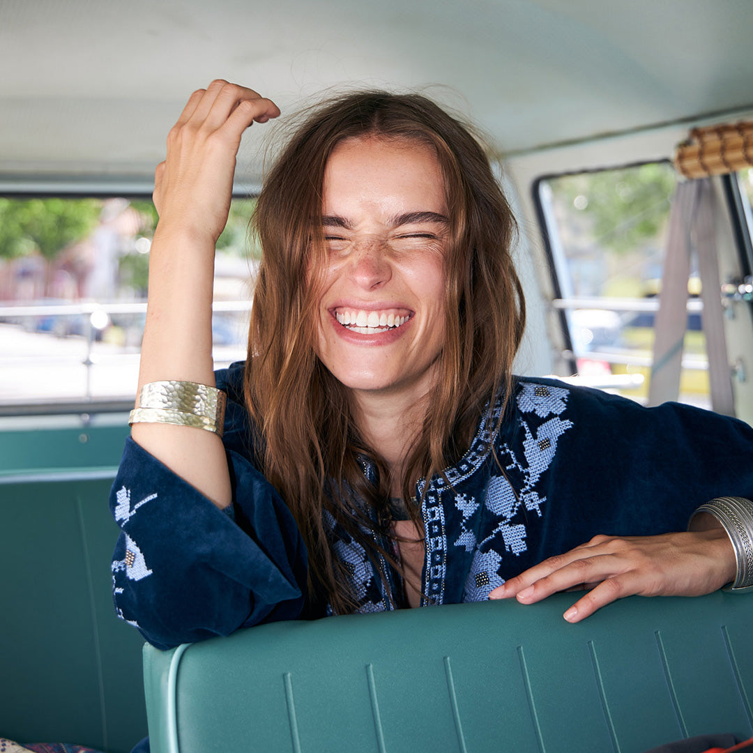 Woman wearing a blue embroidered JACKET sitting in a car, smiling with her eyes closed. She has loose hair and is wearing wide bracelets, creating a light and relaxed mood.