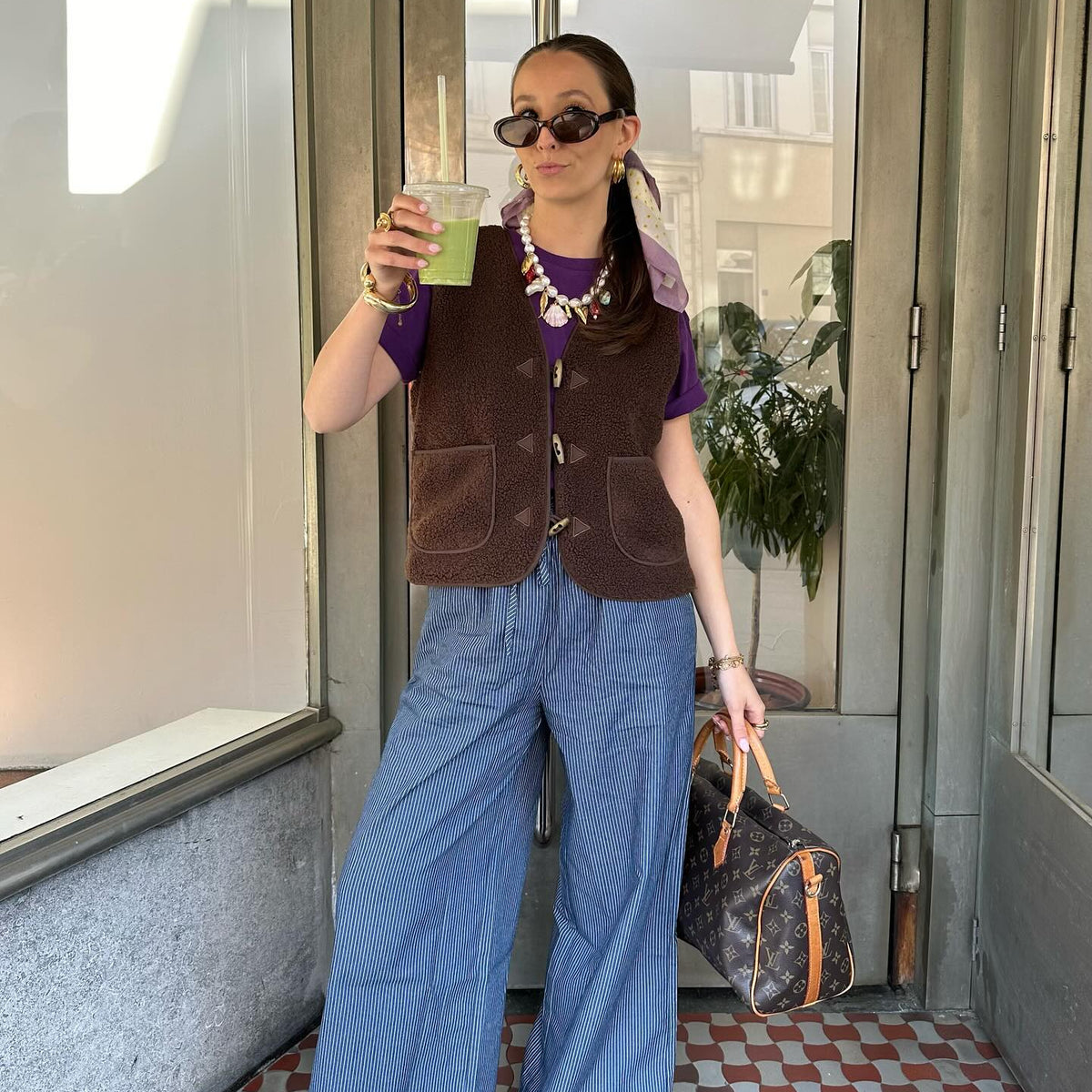 Woman wearing a colourful knitted top with horizontal stripes, styled with white trousers, sunglasses and a black bag in a sunny city square.